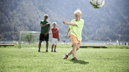 Junge schießt Fußball auf Rasenplatz mit zwei Personen und Tor im Hintergrund