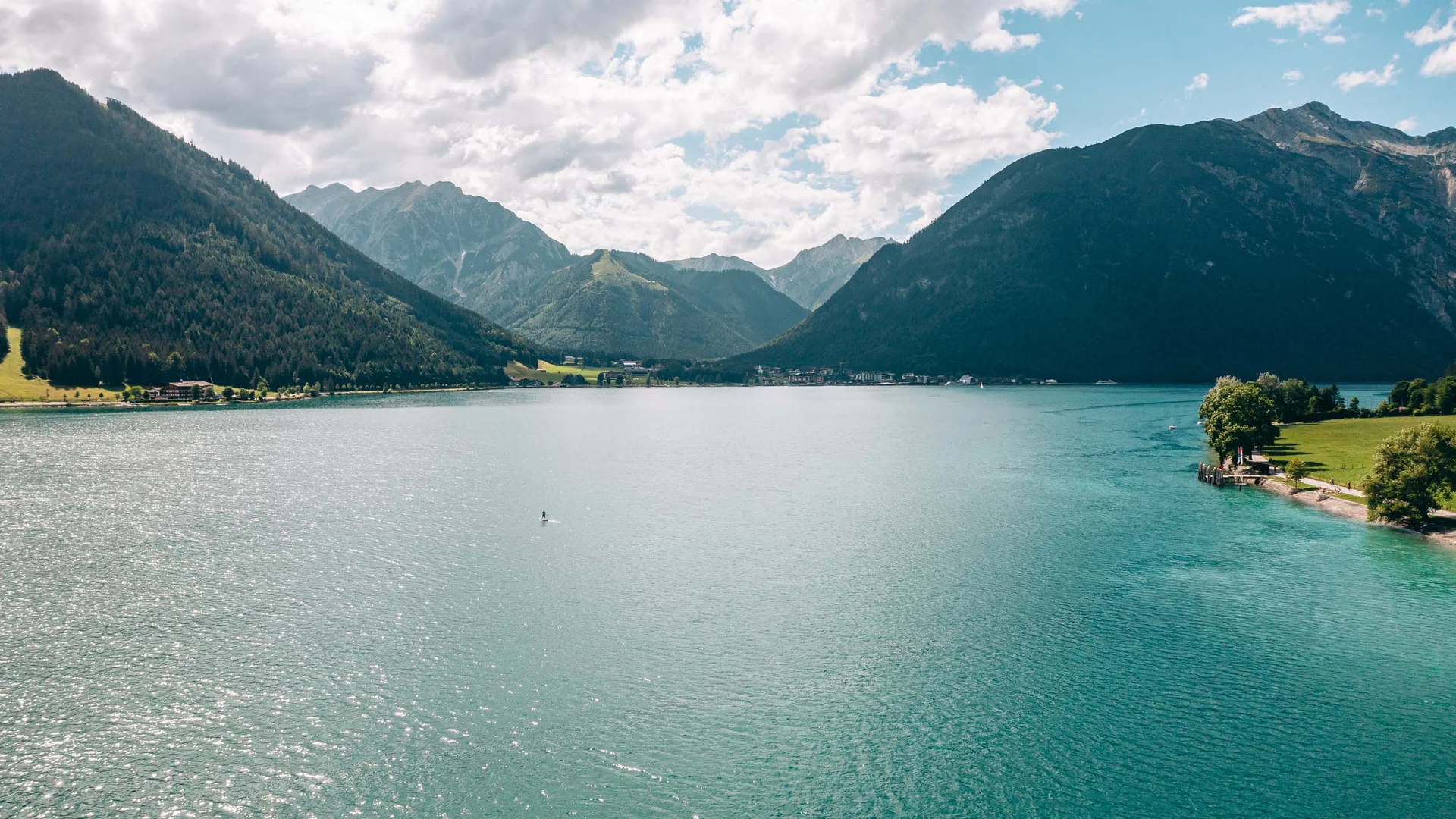 Blick auf einen ruhigen Bergsee mit bewaldeten Hügeln und bewölktem Himmel
