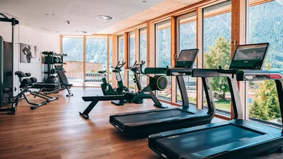 Fitnessraum mit Laufbändern, Fahrrädern und Gewichten vor großen Fenstern mit Bergblick