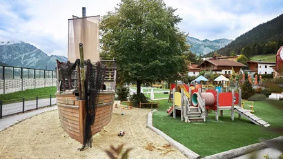 Spielplatz mit Piratenschiff, Sandbereich und buntem Klettergerüst vor Bergkulisse