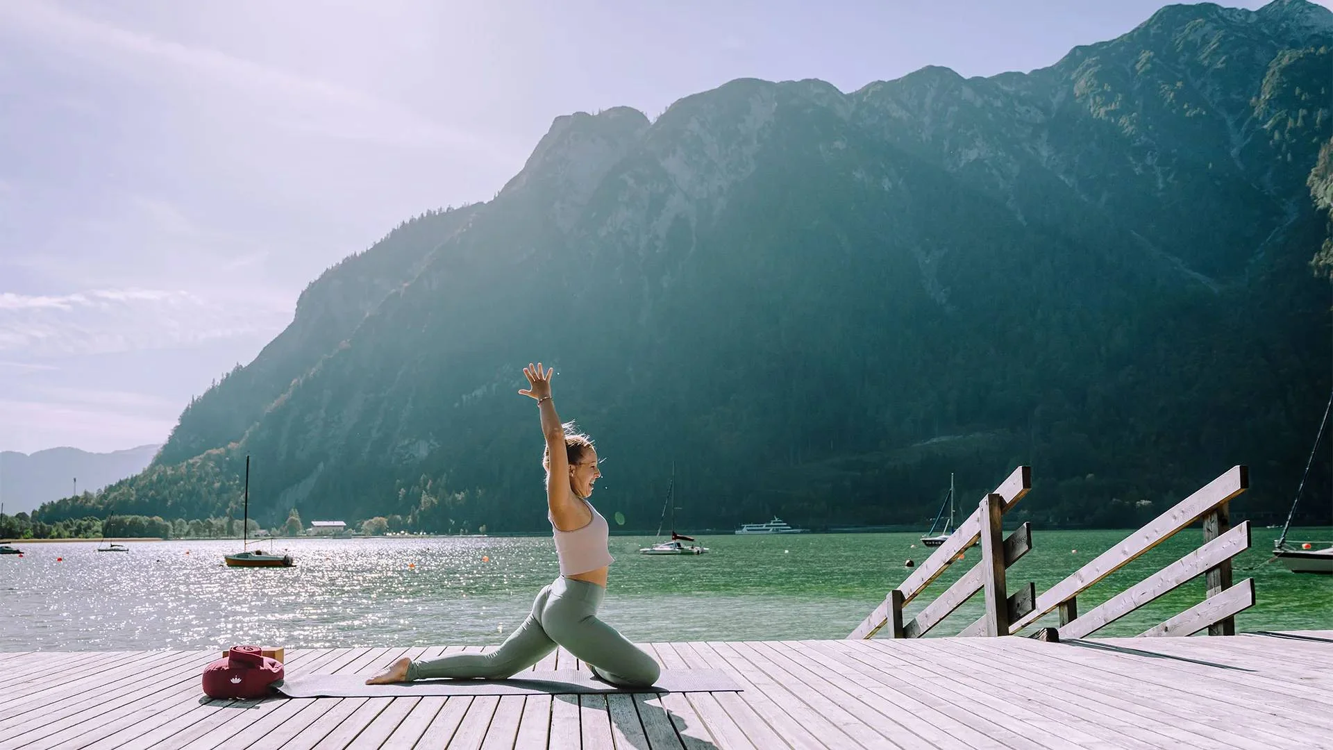 Frau macht Yoga auf Steg vor Bergsee mit Segelbooten