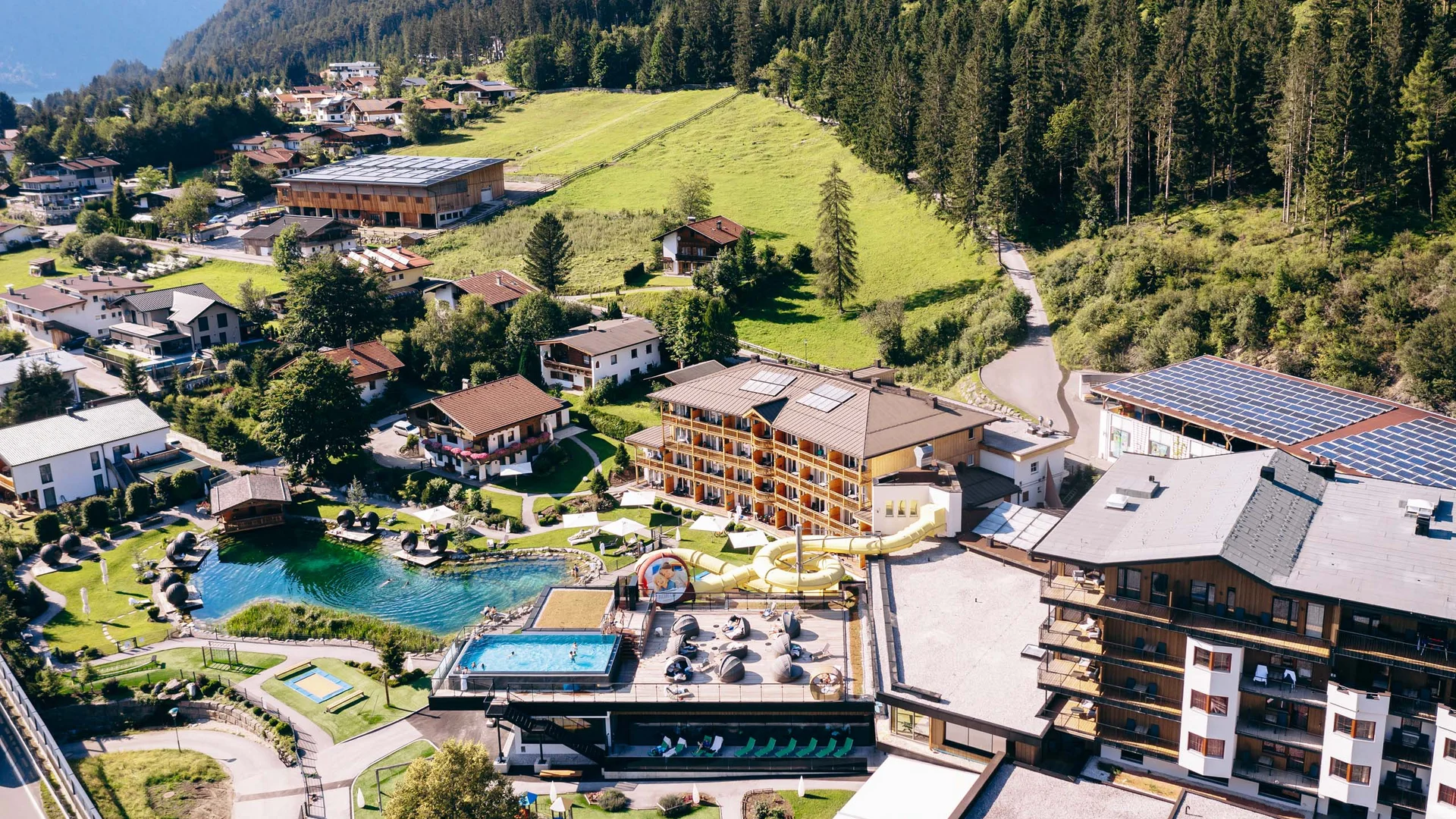 Luftaufnahme eines Hotels mit Pool, Wasserrutsche und Teich in einer grünen Berglandschaft