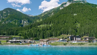 Blick auf blauen See mit Dock, Häusern und bewaldeten Bergen bei klarem Himmel