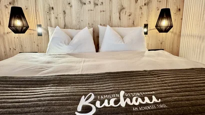 Gemütliches Bett mit weißer Bettwäsche und Buchau Resort Decke vor Holzwand